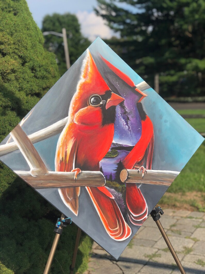 以下が含まれることがあります： 枝に止まっている2羽の赤いカージナルを描いた絵画。カージナルはリアルなスタイルで描かれており、羽毛のディテールや鮮やかな色が表現されています。絵画の背景は濃い青色で、星々の銀河と鳥を通して見える風景が描かれています。