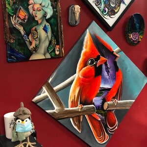 以下が含まれることがあります： 枝に止まっている2羽の赤い鳥の絵。左側の鳥は赤い体と黒い嘴、右側の鳥は紫色の体と黒い嘴をしている。背景は青と白の抽象的なデザインである。