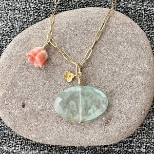 May include: A gold chain necklace with a light green oval pendant and a pink flower charm. The necklace is displayed on a gray stone.