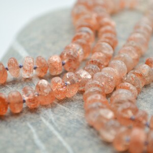 May include: A strand of sunstone beads, with a light orange color and a shimmery, iridescent finish. The beads are strung on a thin, purple cord.