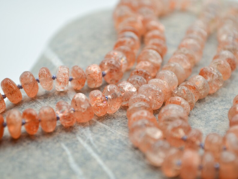 May include: A strand of sunstone beads, with a light orange color and a shimmery, iridescent finish. The beads are strung on a thin, purple cord.