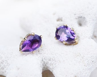 May include: A pair of gold-colored stud earrings, each featuring a pear-shaped, deep purple gemstone. The gemstones are set in a gold-colored metal with four prongs. The earrings are displayed on a white, textured surface.