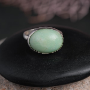 May include: A silver ring featuring a large, oval-shaped, pale green gemstone. The gemstone is set in a simple, polished silver band. The ring is displayed on a dark, textured surface, creating a contrast with the light-colored stone.