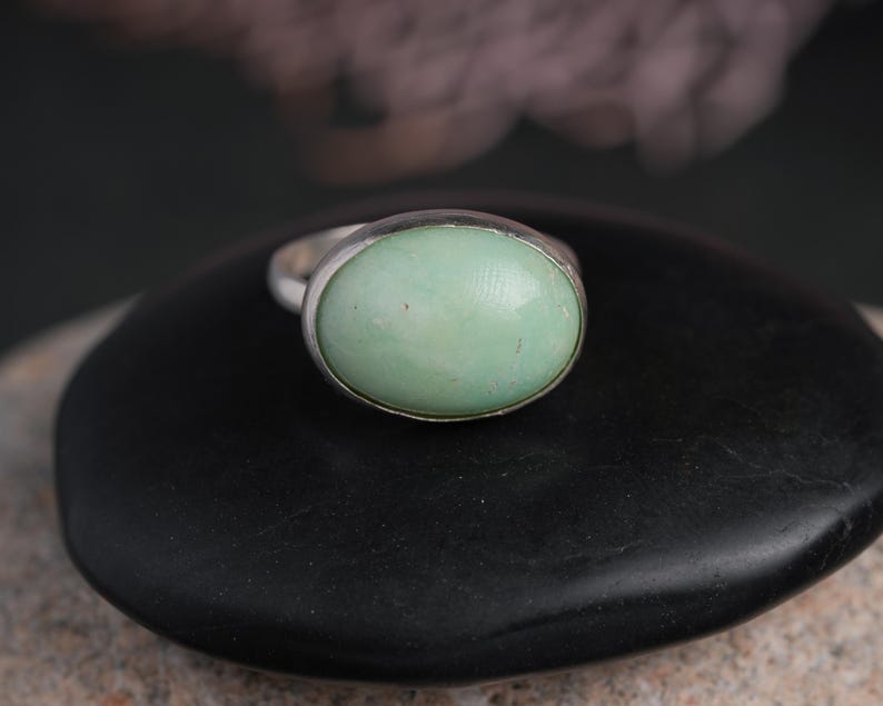 May include: A silver ring featuring a large, oval-shaped, pale green gemstone. The gemstone is set in a simple, polished silver band. The ring is displayed on a dark, textured surface, creating a contrast with the light-colored stone.