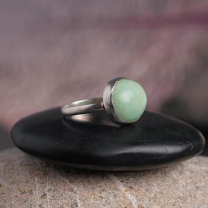 May include: A silver ring featuring a large, round, pale green gemstone. The ring is set on a black, smooth stone, with a blurred background. The gemstone is the focal point, showcasing its soft color and rounded shape. The ring's band is simple and elegant.