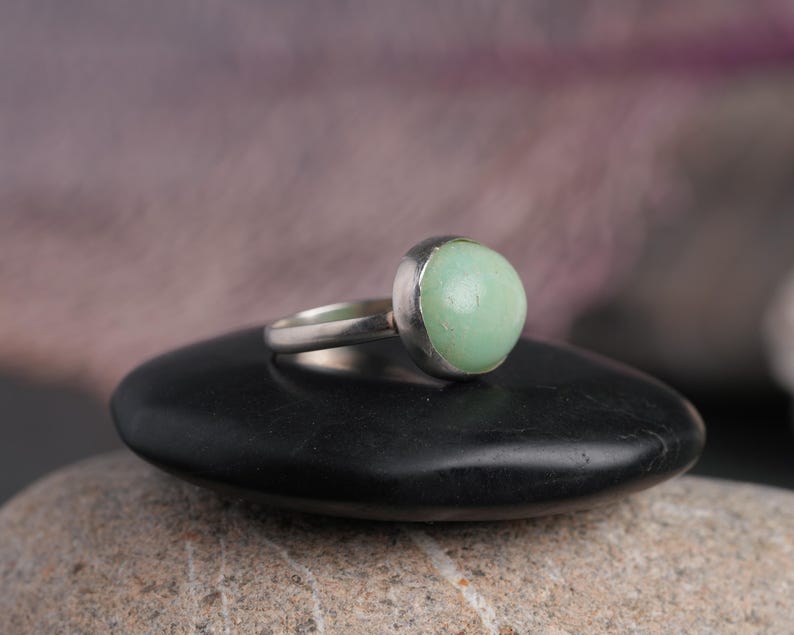 May include: A silver ring featuring a large, round, pale green gemstone. The ring is set on a black, smooth stone, with a blurred background. The gemstone is the focal point, showcasing its soft color and rounded shape. The ring's band is simple and elegant.