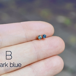 Puede incluir: Un par de peque&ntilde;os pendientes de piedras preciosas azul oscuro con engastes de oro. Los pendientes se muestran en la mano de una persona. El texto "B dark blue" es visible debajo de los pendientes.