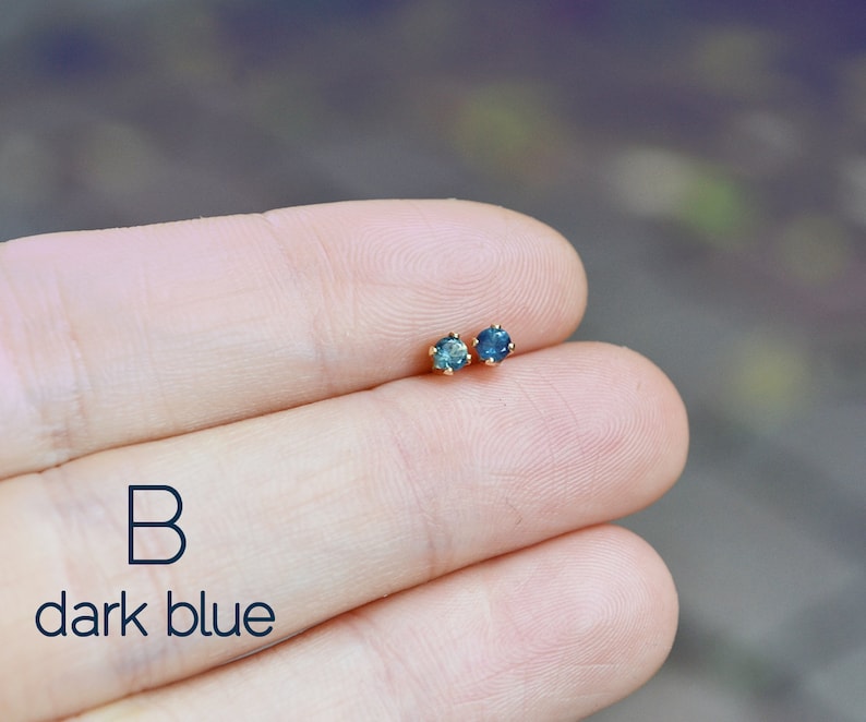 Puede incluir: Un par de peque&ntilde;os pendientes de piedras preciosas azul oscuro con engastes de oro. Los pendientes se muestran en la mano de una persona. El texto "B dark blue" es visible debajo de los pendientes.