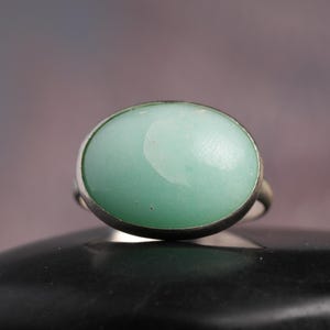 May include: A silver ring featuring a large, oval-shaped, light green gemstone. The gemstone has a smooth, polished surface and is set in a simple silver band. The ring is displayed on a dark surface.