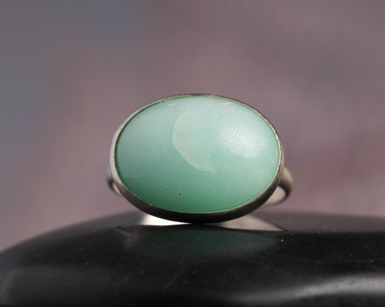 May include: A silver ring featuring a large, oval-shaped, light green gemstone. The gemstone has a smooth, polished surface and is set in a simple silver band. The ring is displayed on a dark surface.