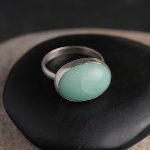 May include: A silver ring with a large, oval, light green stone. The ring is set on a black, rounded surface. The stone has a smooth, polished appearance. The ring's band is simple and thin.