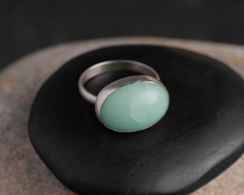 May include: A silver ring with a large, oval, light green stone. The ring is set on a black, rounded surface. The stone has a smooth, polished appearance. The ring's band is simple and thin.