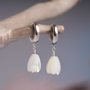 May include: A pair of silver hoop earrings with white, flower-shaped pendants. The earrings are suspended from the hoops by a silver wire. The background is a soft, blurred mix of colors.
