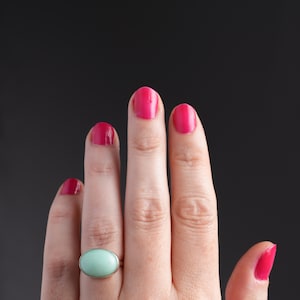 May include: A hand with bright pink nail polish and a silver ring with a light green oval stone. The ring is the focal point, set against a dark background.