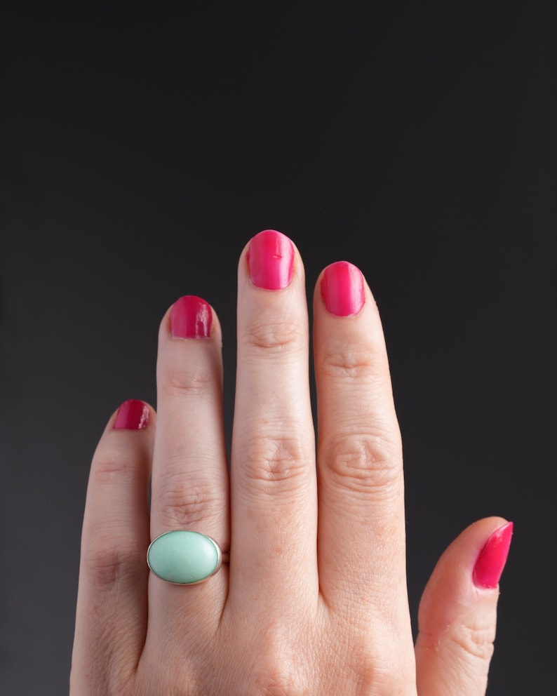 May include: A hand with bright pink nail polish and a silver ring with a light green oval stone. The ring is the focal point, set against a dark background.