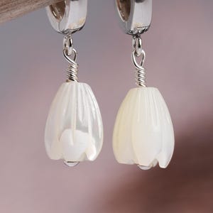 May include: A pair of dangle earrings featuring white, tulip-shaped pendants. The pendants are suspended from silver-colored hoops with a coiled wire detail. The earrings have a pearlescent sheen and a drop of water at the bottom.