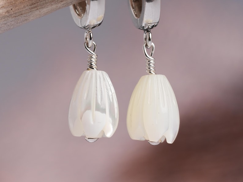 May include: A pair of dangle earrings featuring white, tulip-shaped pendants. The pendants are suspended from silver-colored hoops with a coiled wire detail. The earrings have a pearlescent sheen and a drop of water at the bottom.