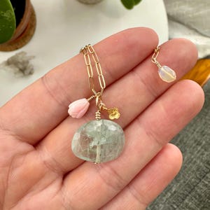 May include: A gold chain necklace with a large, light green aquamarine stone pendant. The pendant is surrounded by smaller charms, including a pink flower and a gold leaf.