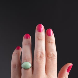 May include: A silver ring with a large, oval, light green stone. The ring is on a hand with bright pink nail polish. The background is a solid black.