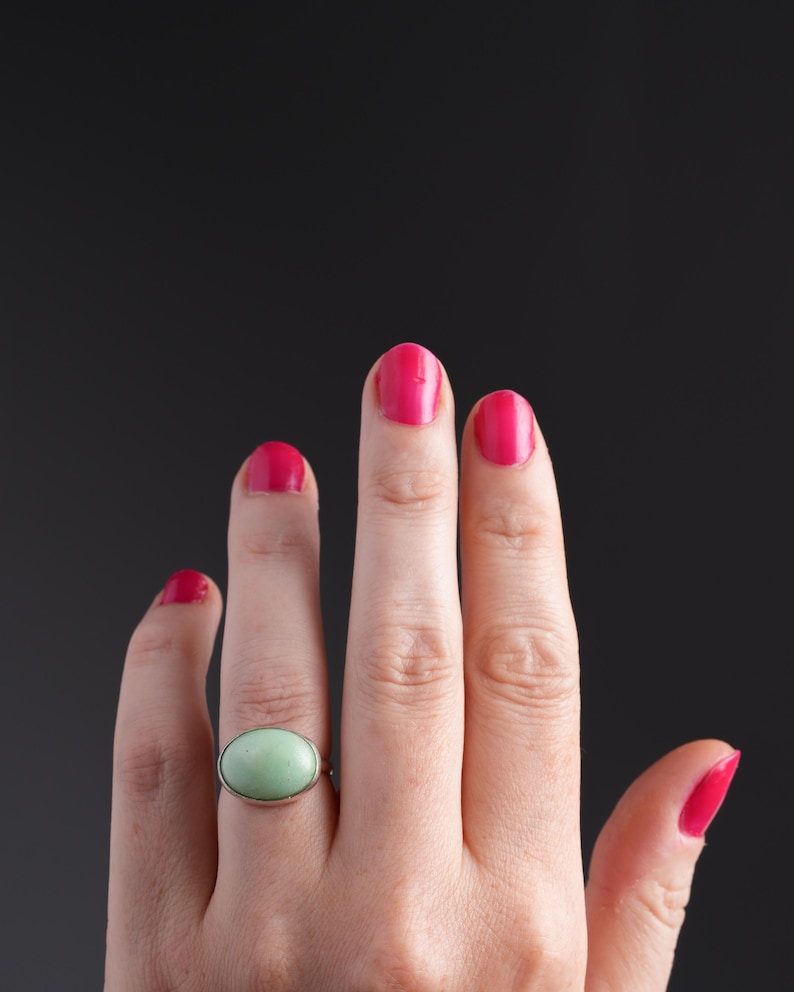 May include: A silver ring with a large, oval, light green stone. The ring is on a hand with bright pink nail polish. The background is a solid black.