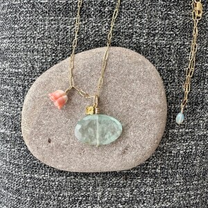 May include: A delicate gold chain necklace with a light blue oval pendant and a pink flower charm. The necklace is displayed on a gray stone.