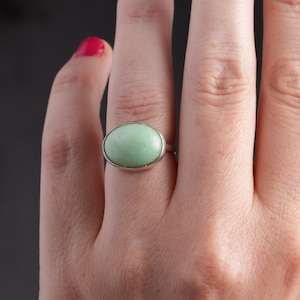May include: A silver ring featuring a large, oval-shaped, pale green stone. The ring is set in a simple, polished silver band. The background is a dark gray.