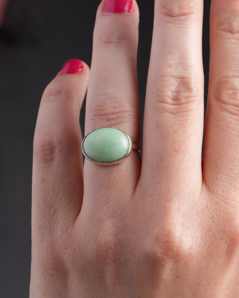 May include: A silver ring featuring a large, oval-shaped, pale green stone. The ring is set in a simple, polished silver band. The background is a dark gray.