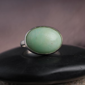 May include: A silver ring featuring a large, oval-shaped, pale green stone. The stone has a smooth, polished surface and is set in a simple silver band. The ring is displayed on a black, rounded surface.