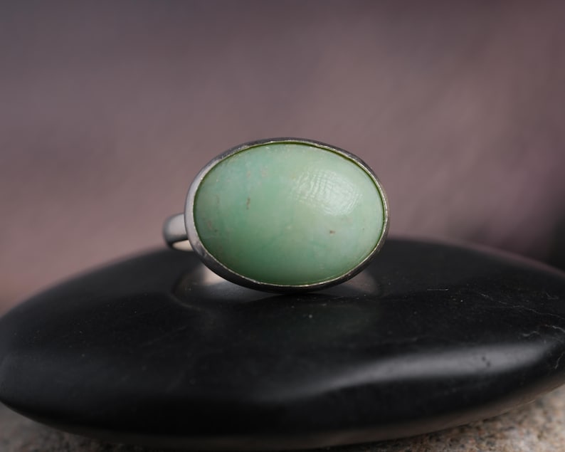 May include: A silver ring featuring a large, oval-shaped, pale green stone. The stone has a smooth, polished surface and is set in a simple silver band. The ring is displayed on a black, rounded surface.
