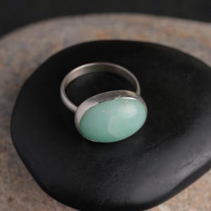 May include: A silver ring with a large, oval, light green stone. The ring's band is thin and metallic, and the stone is set in a bezel setting. The ring is displayed on a black, rounded stone.
