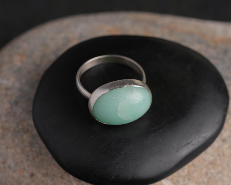May include: A silver ring with a large, oval, light green stone. The ring's band is thin and metallic, and the stone is set in a bezel setting. The ring is displayed on a black, rounded stone.