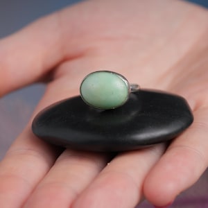 May include: A silver ring with a light green oval stone, resting on a smooth, black, oval-shaped stone. The ring is displayed in the palm of a hand, with the background slightly blurred.