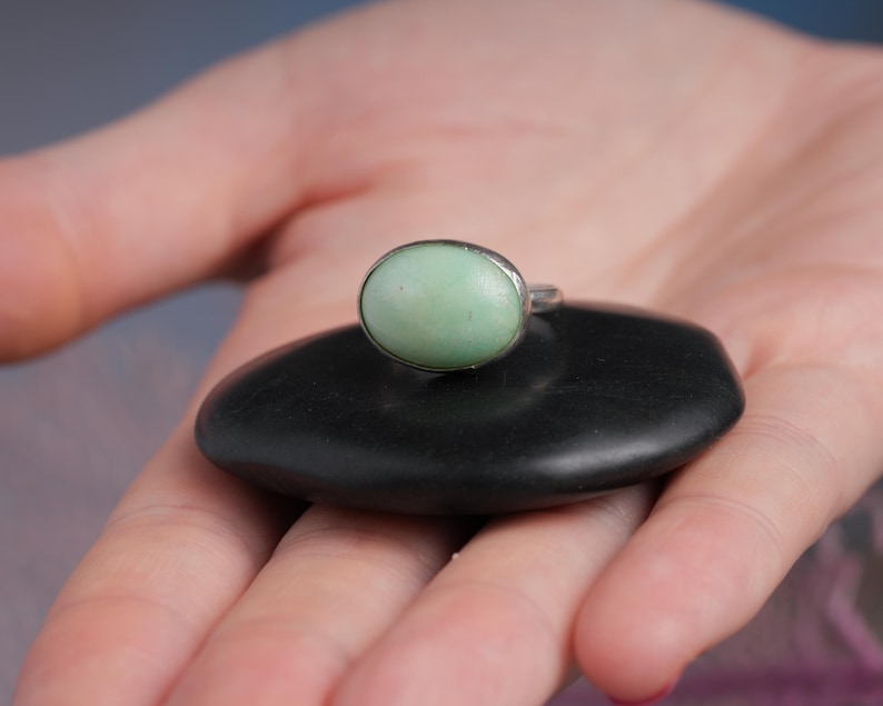 May include: A silver ring with a light green oval stone, resting on a smooth, black, oval-shaped stone. The ring is displayed in the palm of a hand, with the background slightly blurred.