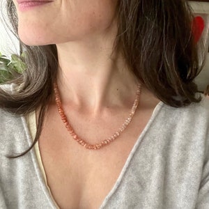 May include: A close-up of a woman wearing a light grey sweater and a necklace made of small, light pink and orange beads.