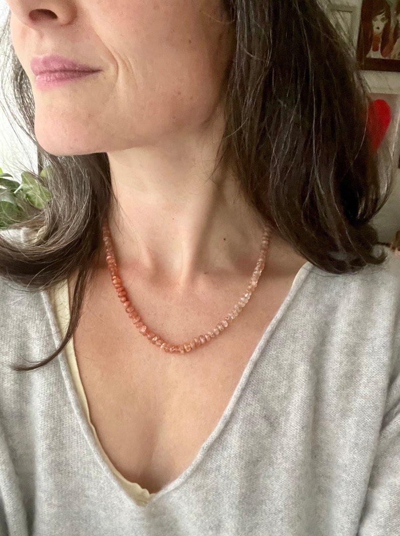 May include: A close-up of a woman wearing a light grey sweater and a necklace made of small, light pink and orange beads.