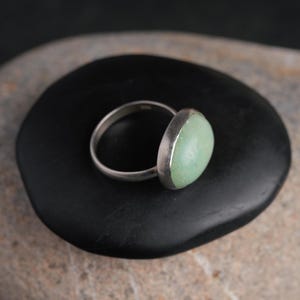 May include: A silver ring with a light green oval stone. The ring is set on a black, smooth stone, with a neutral background. The ring's band is thin and polished, and the stone has a soft, muted color.
