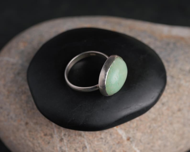 May include: A silver ring with a light green oval stone. The ring is set on a black, smooth stone, with a neutral background. The ring's band is thin and polished, and the stone has a soft, muted color.