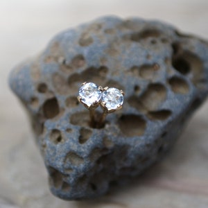 May include: A pair of gold earrings with clear round gemstones. The earrings are on a grey rock.