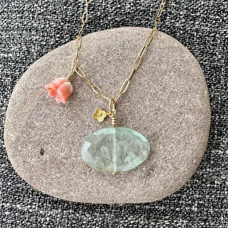 May include: A gold chain necklace with a light green oval pendant and a pink flower charm. The necklace is displayed on a gray stone.