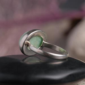 May include: A silver ring with a round, pale green stone. The ring is set in a bezel setting, and the band is simple and elegant. The ring is shown from a side angle, resting on a black surface.