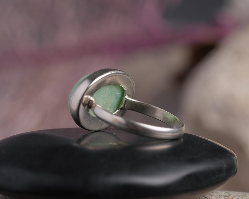 May include: A silver ring with a round, pale green stone. The ring is set in a bezel setting, and the band is simple and elegant. The ring is shown from a side angle, resting on a black surface.