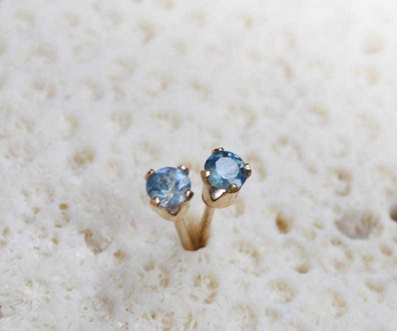 Puede incluir: Un par de pendientes de bot&oacute;n de oro con peque&ntilde;as piedras azules redondas.