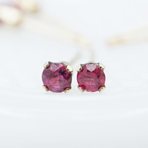 Może przedstawiać: Para małych, okrągłych kolczyków rubinowych z pozłacanymi zapięciami. Kolczyki znajdują się na białym tle.