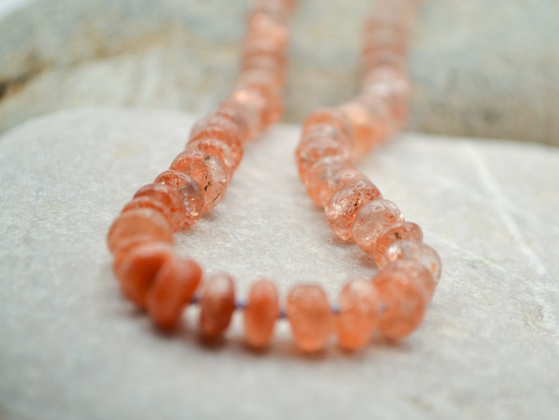 May include: A strand of orange sunstone beads, strung on a clear thread. The beads are faceted and have a translucent appearance.