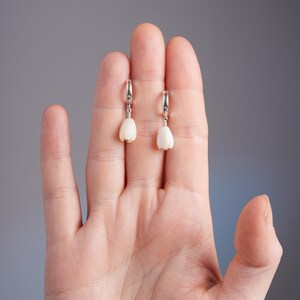May include: A pair of earrings featuring a white, tulip-shaped design suspended from silver hoops. The earrings are displayed against a neutral gray background, showcasing their delicate floral form. These earrings are a stylish accessory.