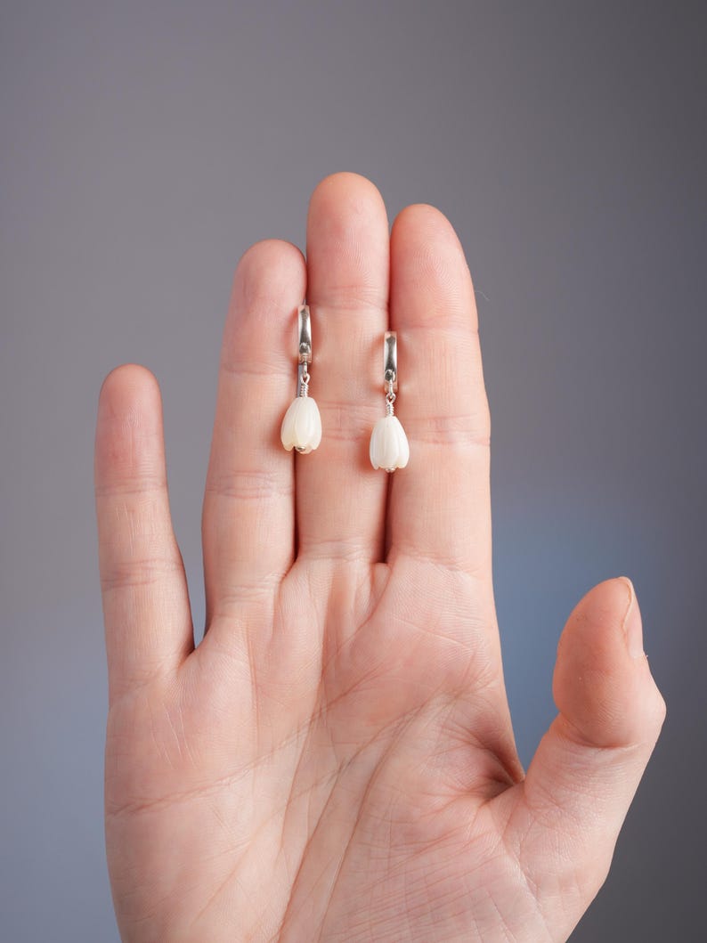 May include: A pair of earrings featuring a white, tulip-shaped design suspended from silver hoops. The earrings are displayed against a neutral gray background, showcasing their delicate floral form. These earrings are a stylish accessory.