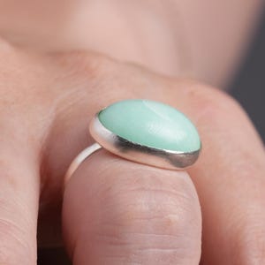 May include: A silver ring featuring a large, oval-shaped, light green stone with a silver border. The ring is worn on a finger, showcasing the polished stone's smooth surface and the ring's simple design.