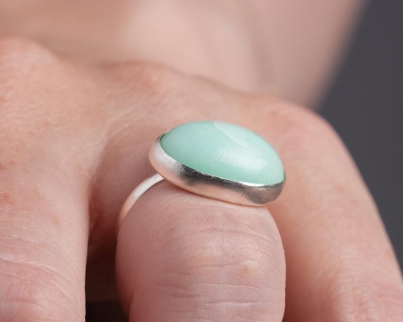 May include: A silver ring featuring a large, oval-shaped, light green stone with a silver border. The ring is worn on a finger, showcasing the polished stone's smooth surface and the ring's simple design.