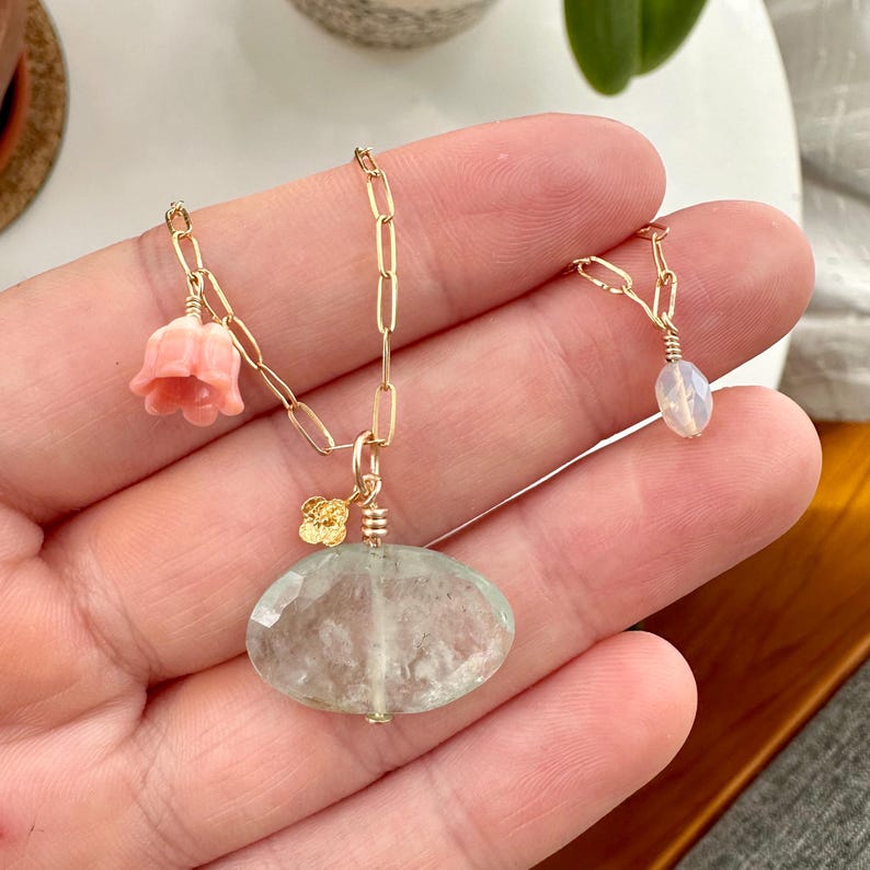May include: A delicate gold chain necklace with a light green oval gemstone pendant and a pink flower charm. The necklace also features a smaller pendant with a white oval gemstone.