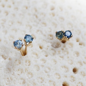 Puede incluir: Un par de pendientes de bot&oacute;n de oro con piedras preciosas azules. Los pendientes est&aacute;n colocados sobre un fondo blanco.